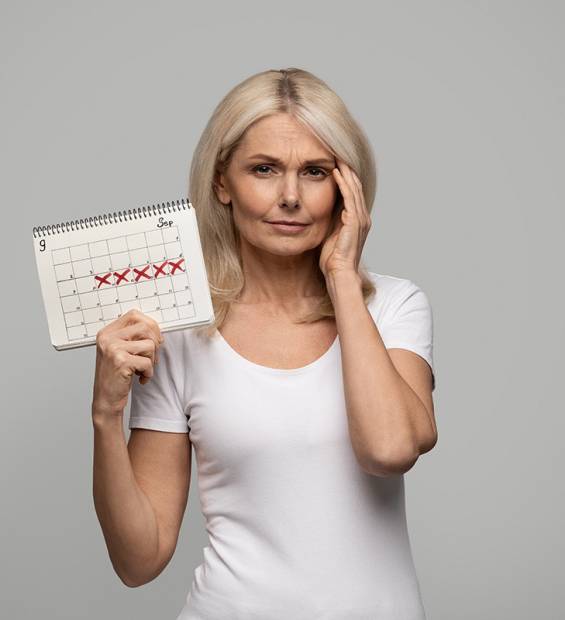 Migraine Tracker. Upset Senior Woman Holding Calendar With Crossed Out Dates And Touching Head, Beautiful Elderly Female Suffering Acute Headache While Standing Over Grey Studio Background